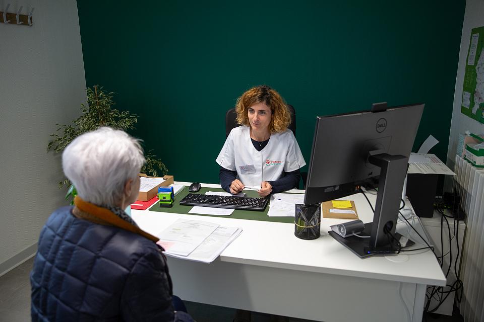 Une femme en blouse blanche assise à un bureau parle à une personne aux cheveux blancs assise en face d'elle.