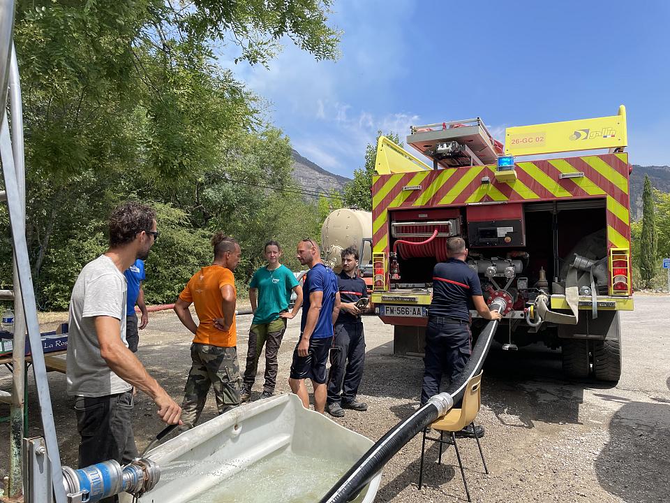 Un camion de pompier tire de l'eau d'une cuve, au milieu d'habitants et de quelques pompiers. 
