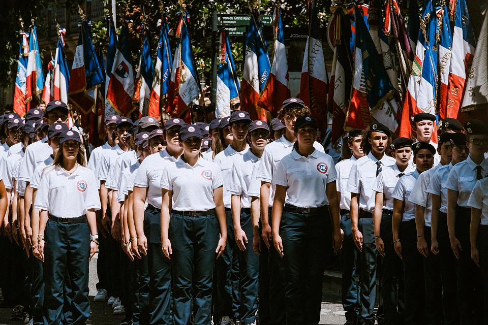 des jeunes, casquettes bleues, tee-shirts blancs, pantalon bleu, en rang et au garde-à-vous devant une rangée de drapeaux français