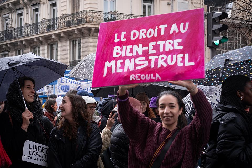 Dans une manifestation, une femme, qui sourit en regardant vers l'objectif, tient au dessus de sa tête une pancarte rose sur laquelle il est écrit en lettres blanches « Le droit au bien être menstruel »