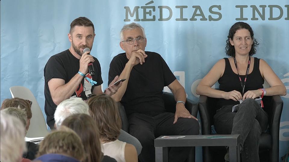 Un homme tient un micro lors d'un débat, deux autres personnes sont assises à côté.