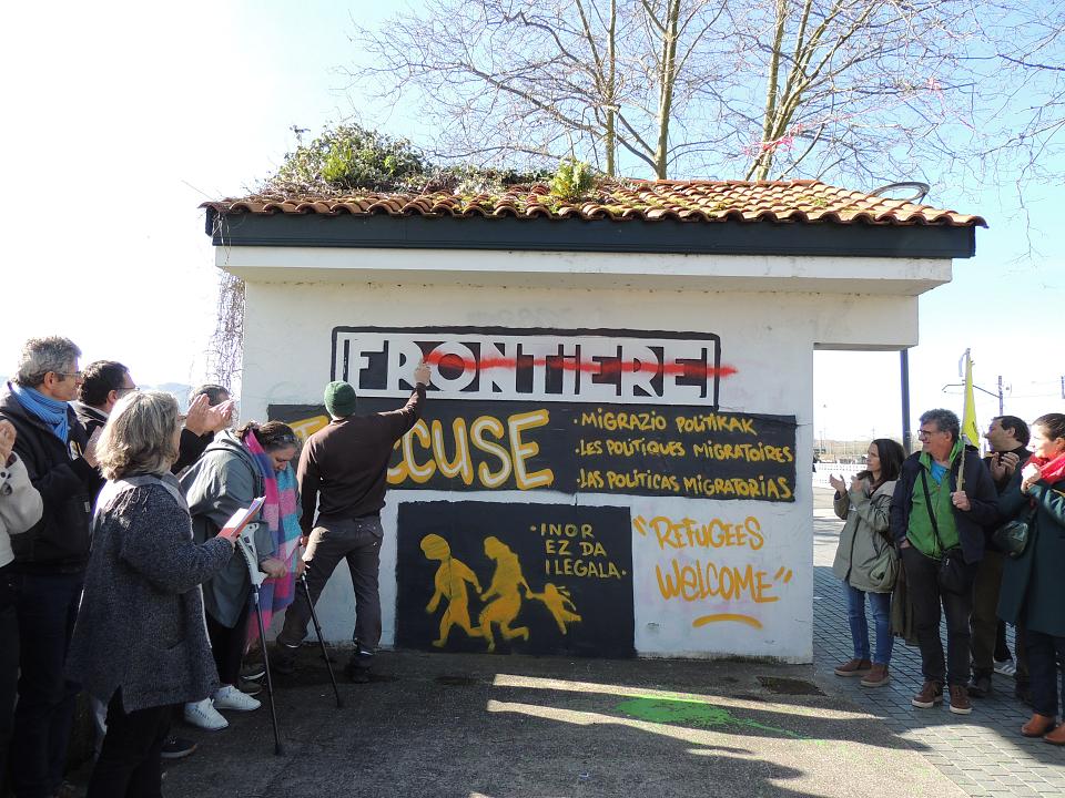 Une vingtaine de personnes réunies autour d'un bloc de béton sur lequel il est écrit "Frontière". Un homme barre le "Frontière" à la peinture de tag rouge. Dessous, on voit écrit en jaune "J'accuse" et l'image d'une famille qui fuit à pied peinte au pochoir.