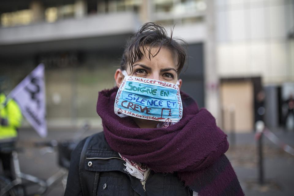 Une femme avec un masque chirurgical sur lequel est écrit "soigne en silence et crève". Dans le fond, une personne tient un drapeau du syndicat Sud. 