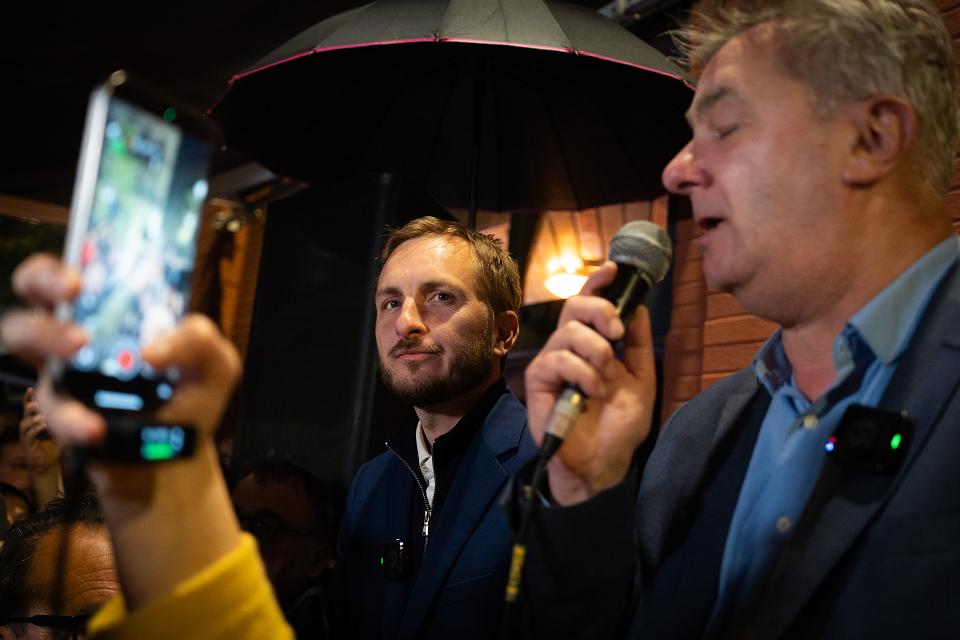 Deux hommes se tiennent côte à côte à l'extérieur, de nuit, sous un grand parapluie noir. L'homme à droite parle dans un micro, les yeux fermés, comme lors d'un discours ou d'une prise de parole publique. L'homme à gauche regarde droit devant lui, calme et attentif, portant une veste sombre avec un petit micro accroché. Au premier plan, une personne filme la scène avec un smartphone.