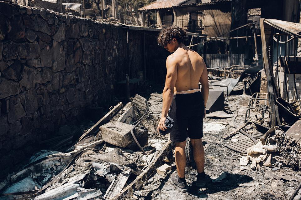 Un jeune homme en short et torse nu regarde au sol les cendres d'objects calcinés dans ce qu'il reste d'une cour ou d'une ruelle. 
