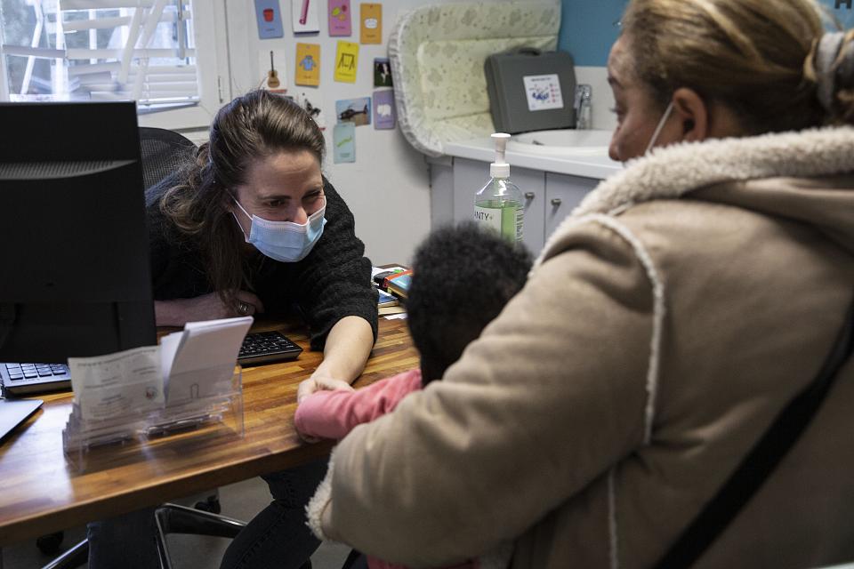 Une femme avec un masque penchée sur un bureau parle à un enfant assis de l'autre côté de la table sur les genou d'une femme. 