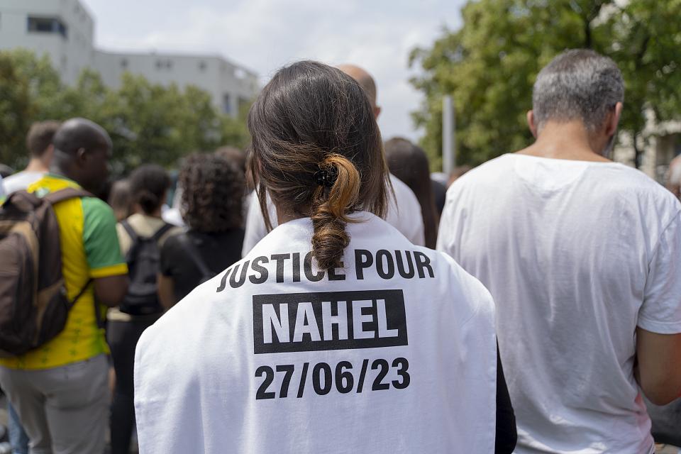 Dans une manifestation, une femme de dos a sur son dos un tissu blanc sur lequel il est écrit "Justice pour Nahel, 27/06/23"