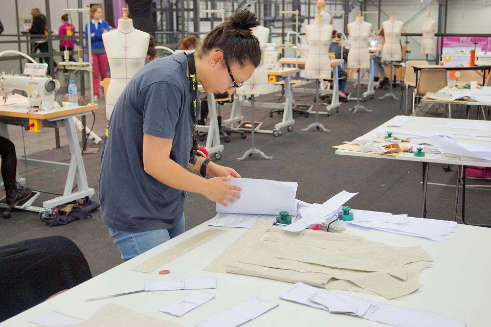 Une jeune est débout penchée sur une table et examine des dossiers. Des mannequins de couture et des tables avec des machines à coudre se trouvent dans la large pièce. 