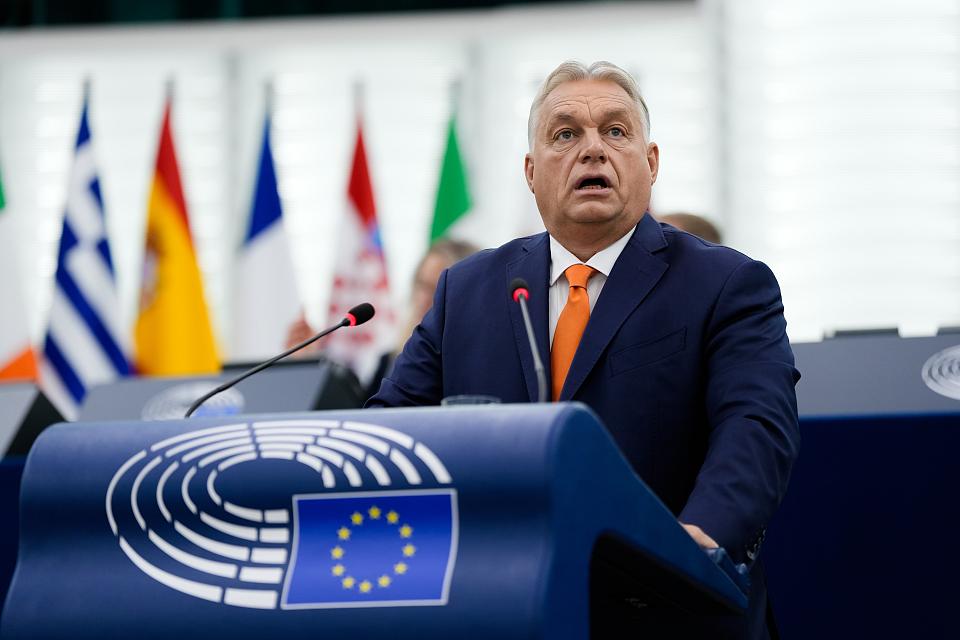 Viktor Orbán parle à un pupitre sur lequel on voit le symbole d'un hémicycle et le drapeau européen. On voit des drapeaux au deuxième plan.