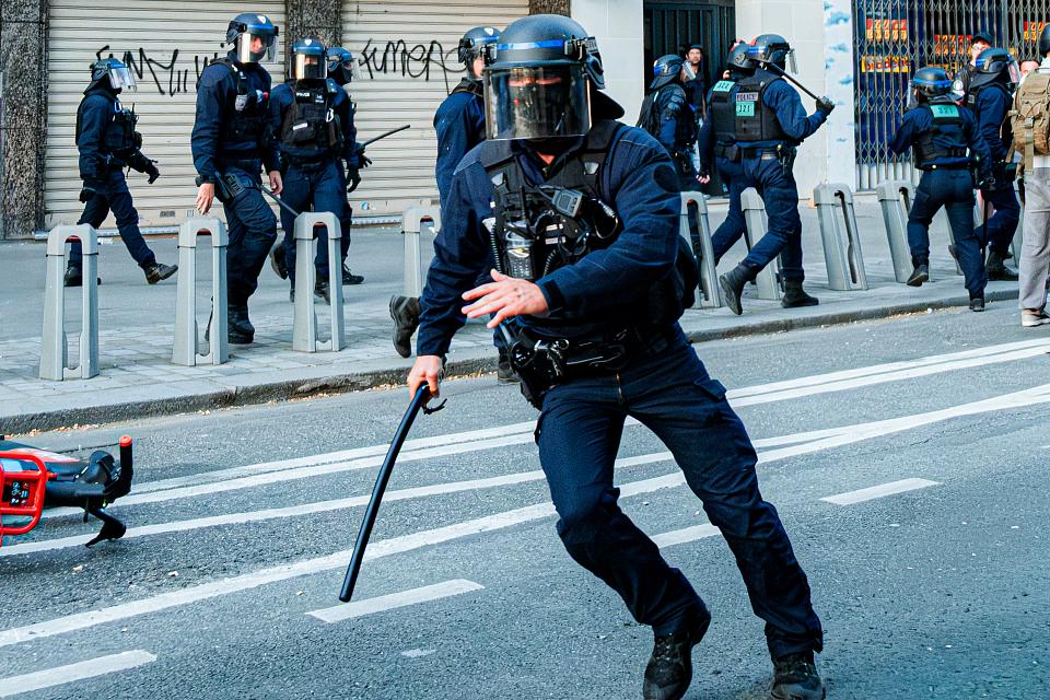 Un policier casqué court avec une matraque à la main dans une rue, d'autres policiers en uniforme de maintient de l'ordre se trouvent au second plan. 