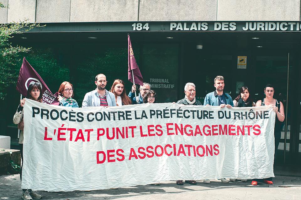 Dix personnes debout derrière une banderole sur laquelle il est écrit « Procès contre la préfecture du Rhône. L'État punit les engagements des associations. » Sur la bâtiment derrière, il est écrit « Palais de justice ».