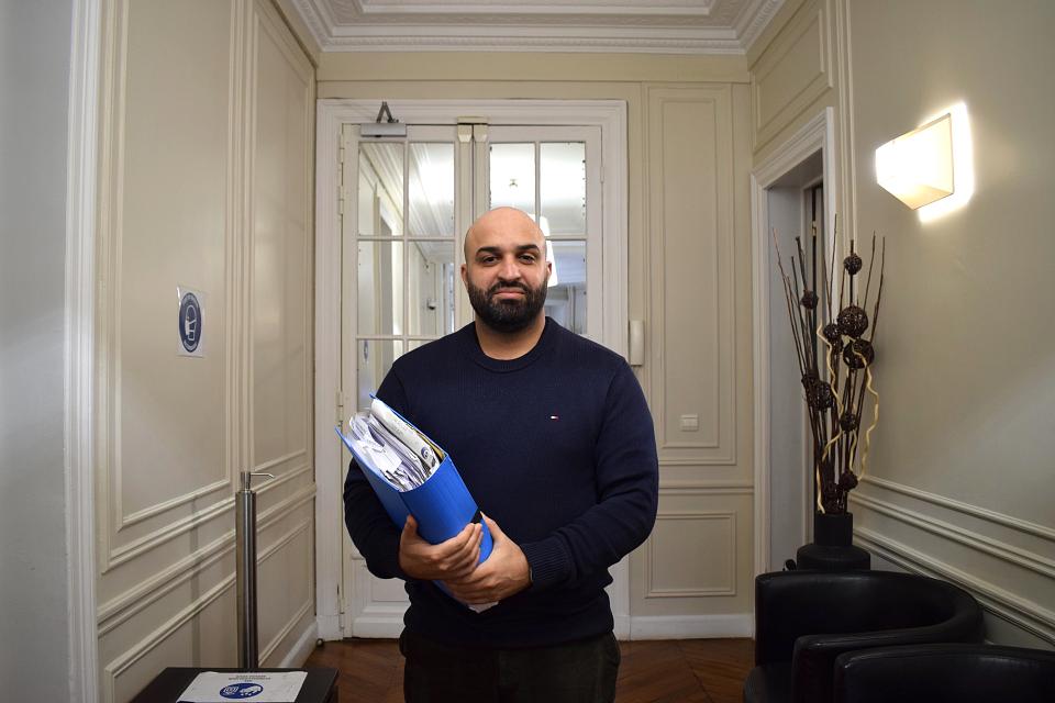 Un homme debout dans un couloir, regarde l'objectif, il porte des dossier dans son bras droit.