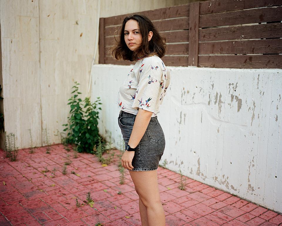 Une jeune femme en short et chemise debout dans une cour regarde l'objectif. Elle ne sourit pas.