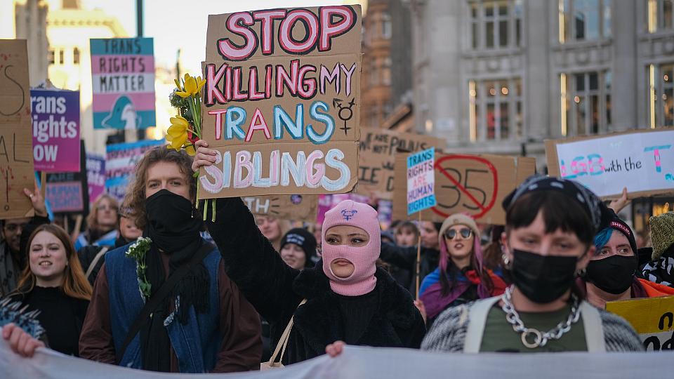 Une personne avec une cagoule rose tient, dans une manifestation, une pancarte avec écrit "Stop killing my trans siblings"