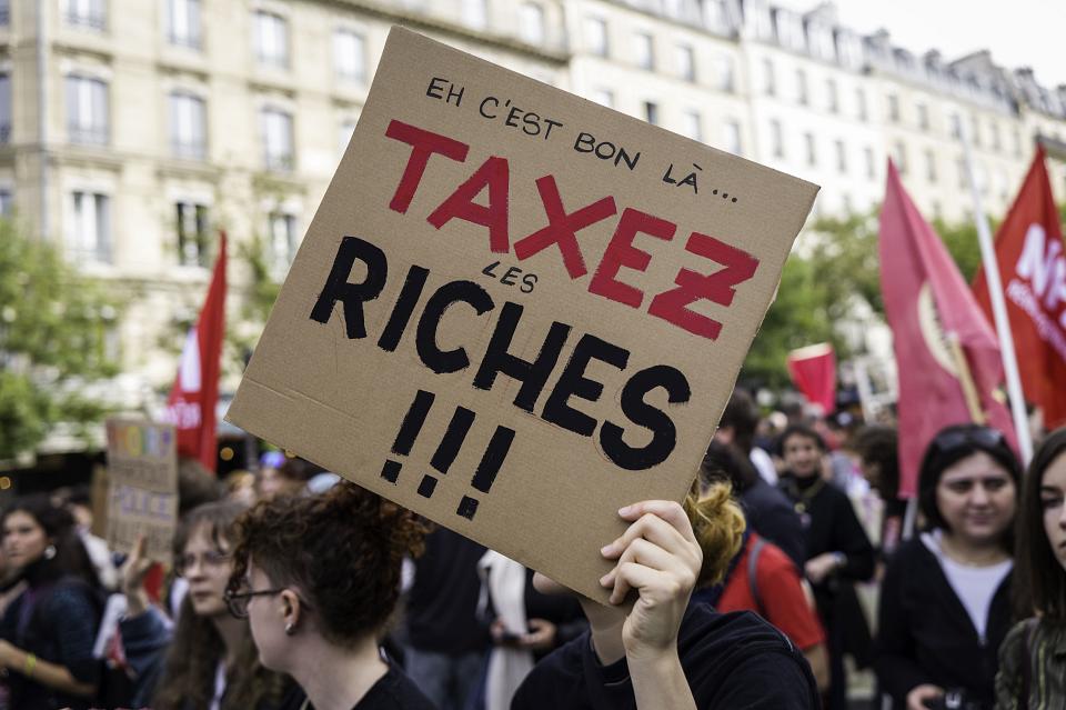 Un manifestant tient une pancarte avec le texte «Eh c'est bon là...Taxer les riches». 