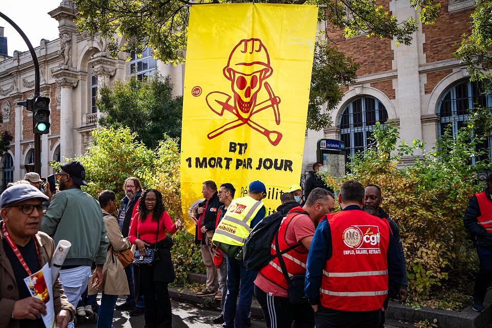Des manifestants dont certains avec un chasuble rouge de la CGT devant une banderole jaune sur laquelle est dessinée en rouge un tête de mort coiffée d'un casque de chantier et est écrit « BTP, un mort par jour ». 
