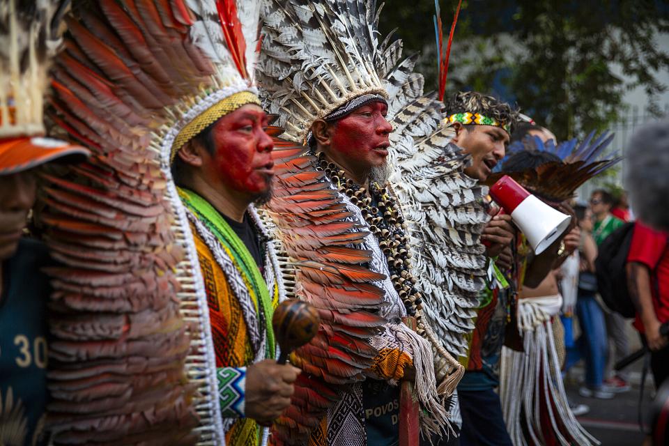 Les visages des quatre hommes de communautés autochtones en tête du cortège d'une manifestation, vus de profil. L'un tient un porte-voix. Deux hommes ont le visage couvert de couleur rouge et porte des coiffes de plumes. 