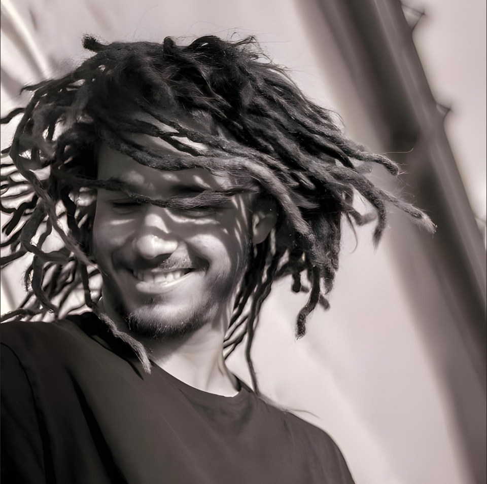 Un jeune homme souriant de profil, avec des dreadlocks et une légère barbe. La photo est en noir et blanc.