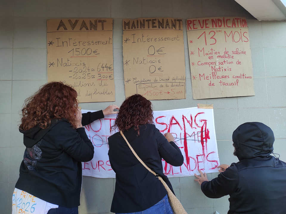 Des revendications inscrites sur des pancartes sont affichées au mur. Trois personnes affichent une banderole en plus. Elles sont photographiées de dos.