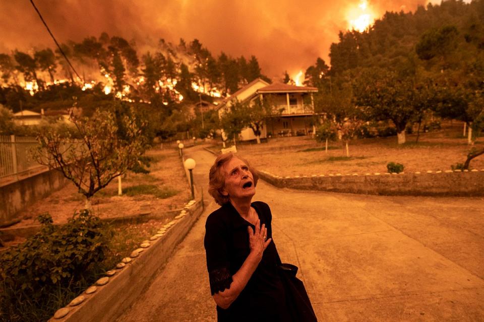 Une femme âgée semble crier devant un paysage en feu qui rougit le ciel. 