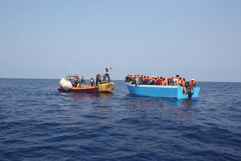 Une embarcation où sont assises un grand nombe de personnes munies de gilets orange face à un canot avec des sauveteurs à bord. 