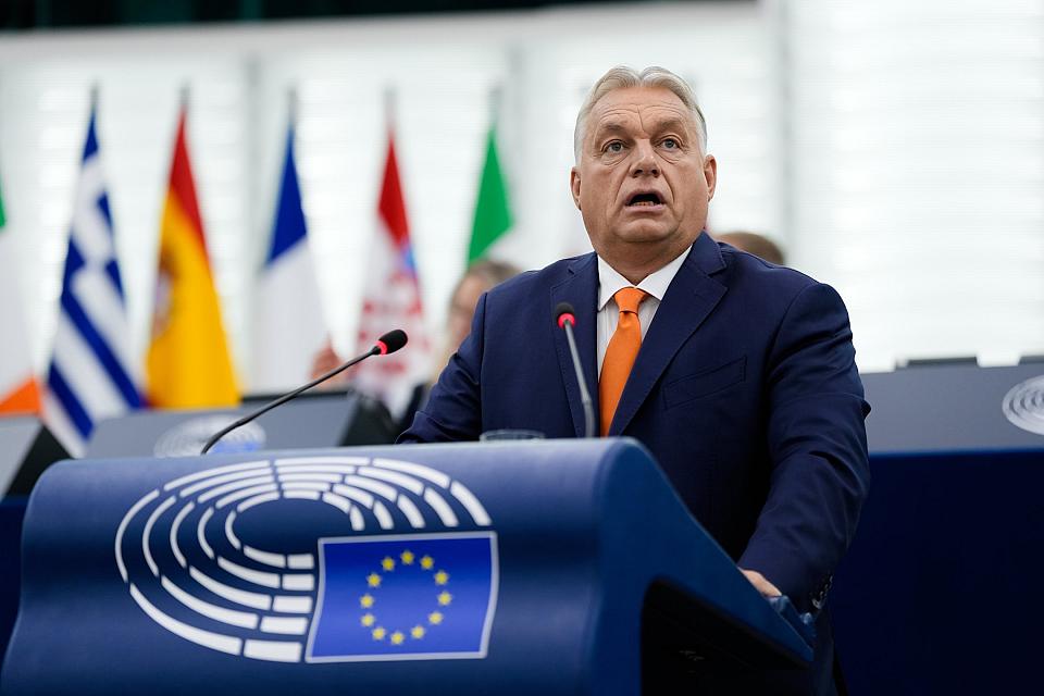 Viktor Orbán parle à un pupitre sur lequel on voit le symbole d'un hémicycle et le drapeau européen. On voit des drapeaux au deuxième plan. 