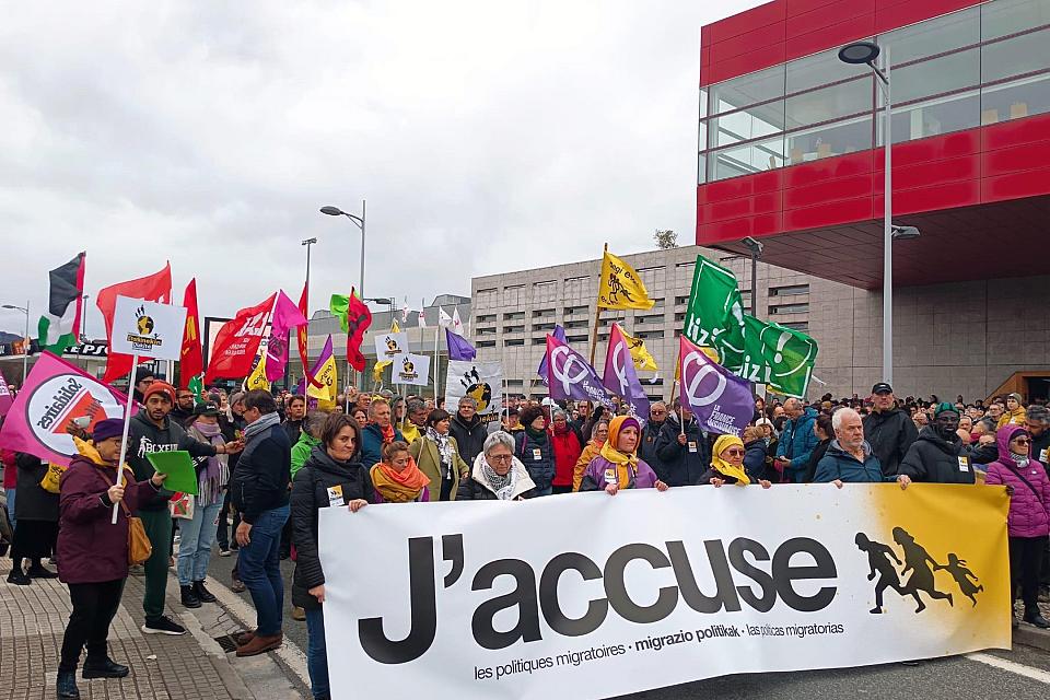 Une banderole "j'accuse" derrière laquelle des dizaines de personnes arborent des drapeaux d'organisations associatives, syndicales et de partis