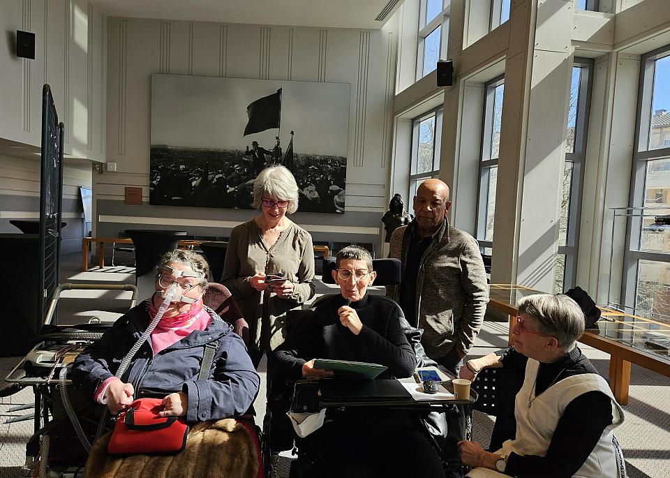 Un groupe de 5 personne se tient dans un espace lumineux. Deux personnes sont assises en fauteuil roulant, d'autres sont debout autour d'elles. En arrière-plan, de grandes fenêtres et une photographie historique accrochée au mur.