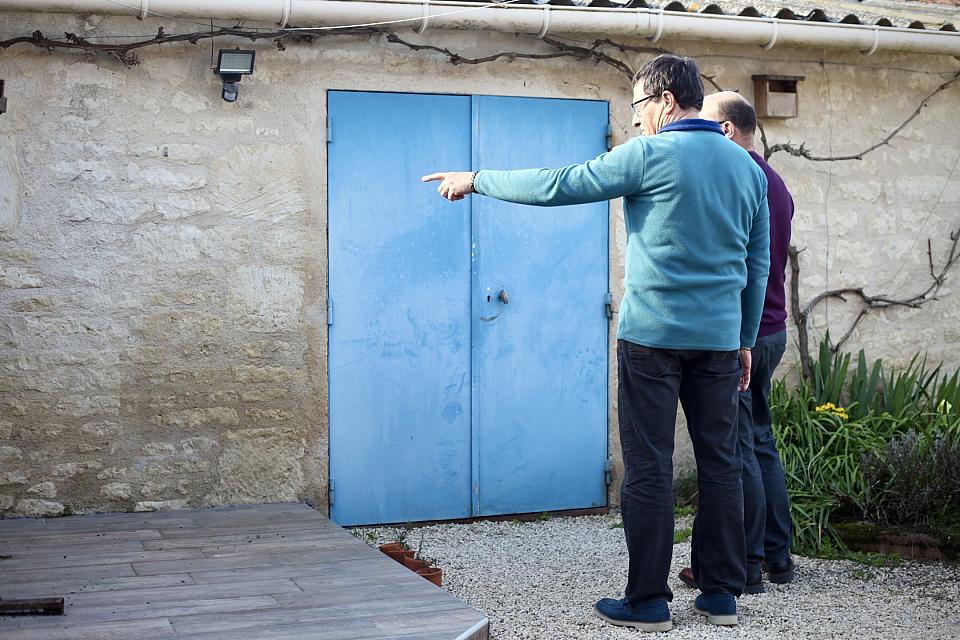 Alain et Hugo*, montrant l'une des façades de leur maison qui a été recouverte de tags homophobes. Devant eux un mur en pierre et une porte bleue