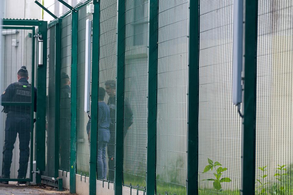 Un policier, derrière une porte grillagée et des personnes derrière un grillage.