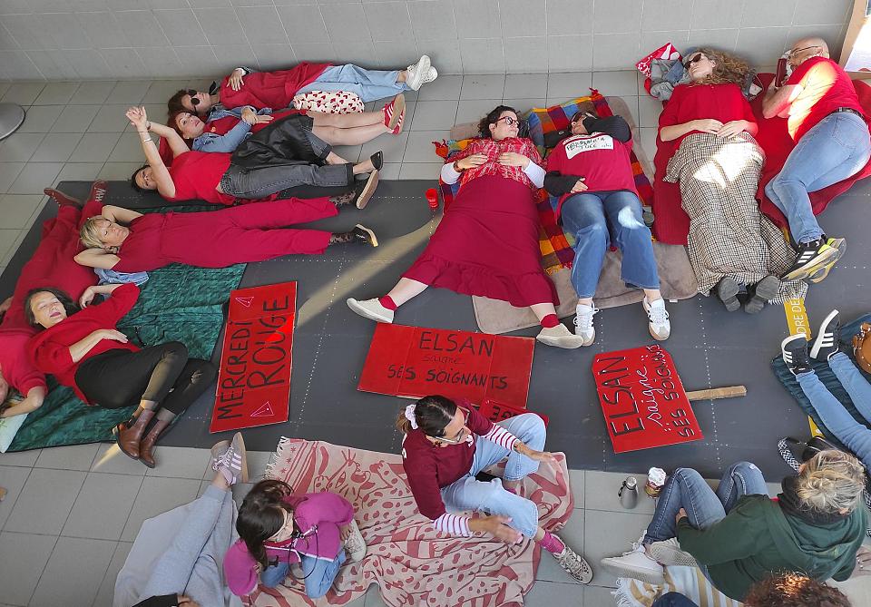 Des personnes vêtues de rouges sont allongées par terre à côtés de pancartes rouges. Sur l'une d'elle on peut lire "Elsan saigne ses soignantes".