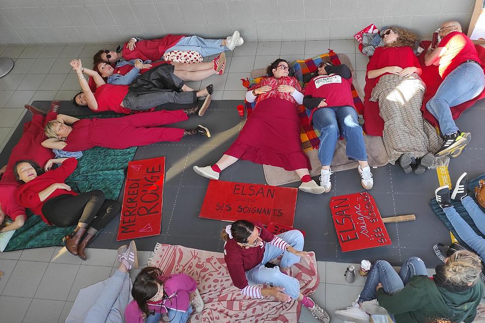 Des personnes vêtues de rouges sont allongées par terre à côtés de pancartes rouges. Sur l'une d'elle on peut lire "Elsan saigne ses soignantes".