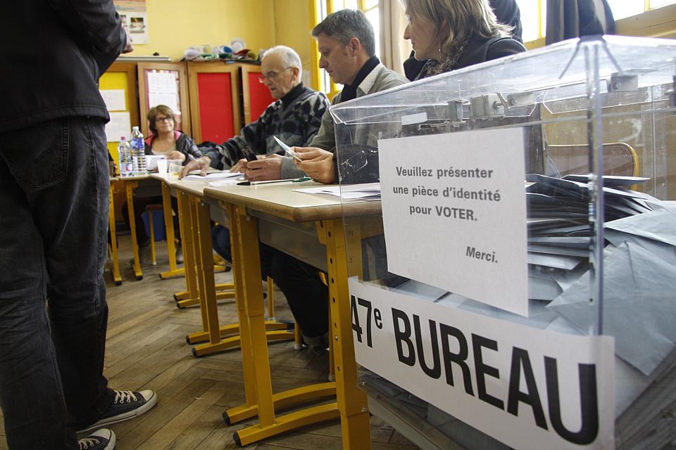 Vue d'un bureau de vote, l'urne au premeir plan, des personnes assises au second plan. 