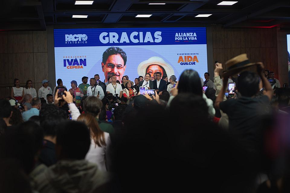 Image d'Ivan Cepeda et de sa co-candidate sur un écran au second plan, une foule de dos dans une salle se trouve au premier plan.