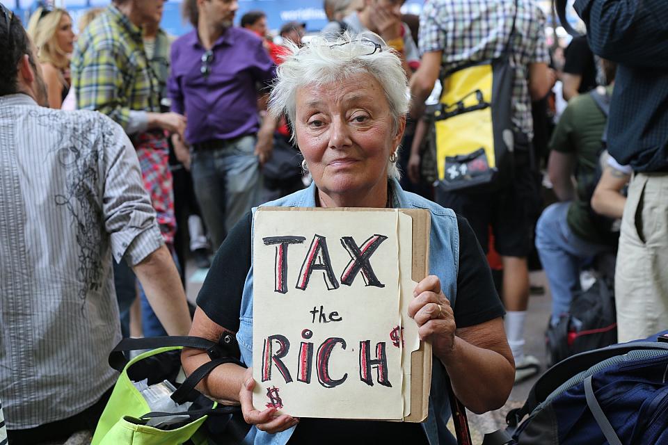 Dans un rassemblement, une femme tient une pancarte sur laquelle il est écrit "Tax the rich". 
