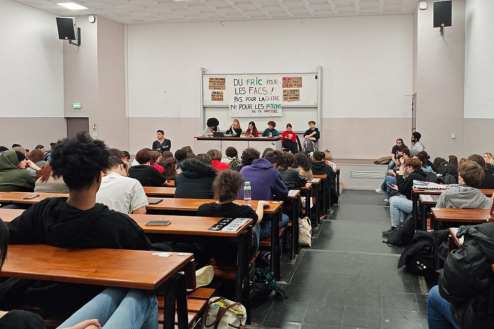 Des étudiants dans une amphi. Cinq jeunes sont assis derrière un bureau sur l'estrade. Une jeune femme parle au micro. Derrière est suspendu une banderole sur laquelle on peut lire « Du fric pour les facs ». 