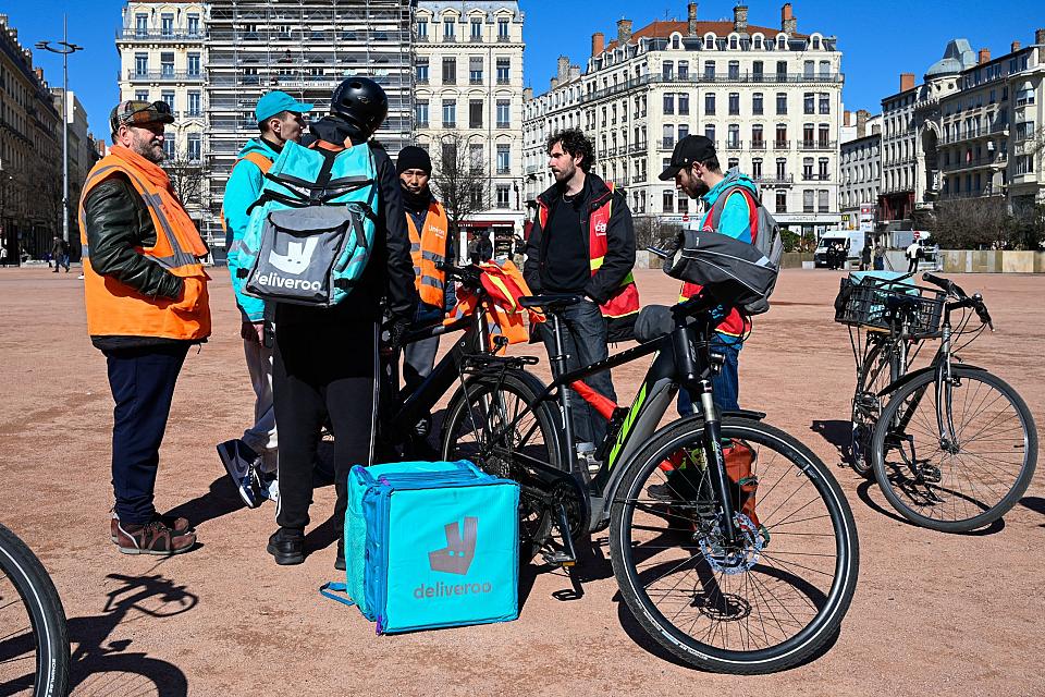 Des livreurs a vélos Uber Eats et Deliveroo portant des gilets CGT et Union indépendants discutent ensemble à côté de vélos et de sacs de livraison. 