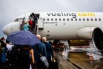 Des passagers vus de dos montant dans un avion sur lequel est écrit Vueling. Le ciel est gris, une femme tient un parapluie.
