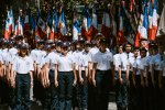 des jeunes, casquettes bleues, tee-shirts blancs, pantalon bleu, en rang et au garde-à-vous devant une rangée de drapeaux français
