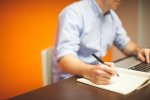 Un homme devant un ordinateur, sa main gauche sur le clavier, sa main droit tient un crayon et semble prendre des notes sur un cahier. On ne voit pas son visage. 