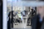 Une femme en uniforme blanc fatiguée au téléphone derrière une porte vitrée avec des reflets dans un couloir d'hôpital.