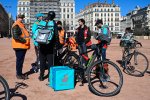 Des livreurs a vélos Uber Eats et Deliveroo portant des gilets CGT et Union indépendants discutent ensemble à côté de vélos et de sacs de livraison. 
