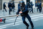 Un policier casqué court avec une matraque à la main dans une rue, d'autres policiers en uniforme de maintient de l'ordre se trouvent au second plan. 