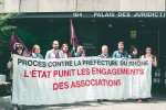 Dix personnes debout derrière une banderole sur laquelle il est écrit « Procès contre la préfecture du Rhône. L'État punit les engagements des associations. » Sur la bâtiment derrière, il est écrit « Palais de justice ».