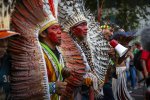 Les visages des quatre hommes de communautés autochtones en tête du cortège d'une manifestation, vus de profil. L'un tient un porte-voix. Deux hommes ont le visage couvert de couleur rouge et porte des coiffes de plumes. 