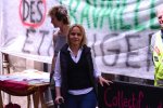 une femme blonde vêtue d'un gilet noir et d'une chemise blanche devant une banderole