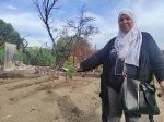 Une femme portant un foulard, un téléphone portable à son cou et un sac à main montre un terrain en friche, brûlé par les flammes.