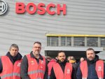 Quatre hommes en chasubles rouge de la CGT, debout devant l'entrée d'une usine sur laquelle est écrit Bosch en lettres rouges, regardent vers l'objectif.