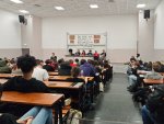 Des étudiants dans une amphi. Cinq jeunes sont assis derrière un bureau sur l'estrade. Une jeune femme parle au micro. Derrière est suspendu une banderole sur laquelle on peut lire « Du fric pour les facs ».