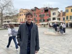Un jeune homme se tient debout, souriant, sur une place dans la ville de Venise.